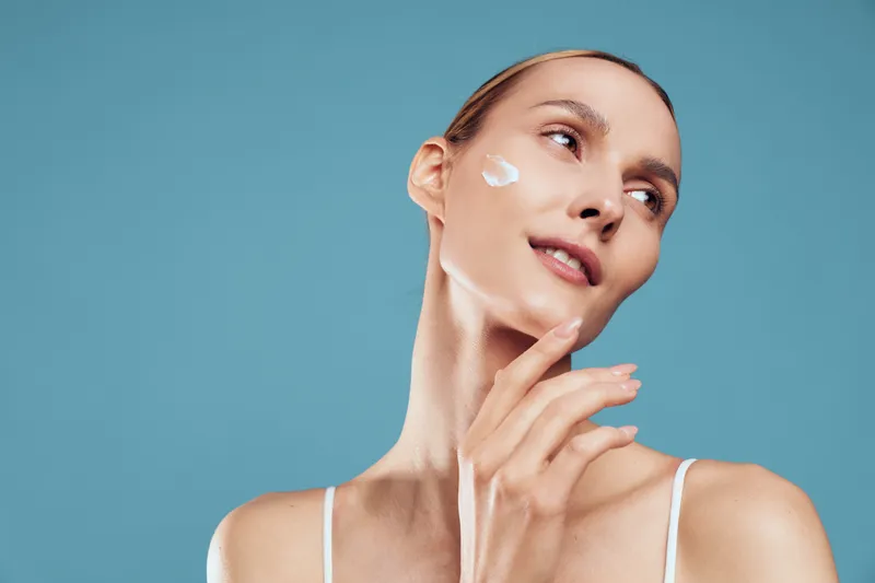 A young woman with clear skin applies cream to her cheek, smiling gently against a blue background. Concept for skincare, beauty routine, and healthy skin care habits.