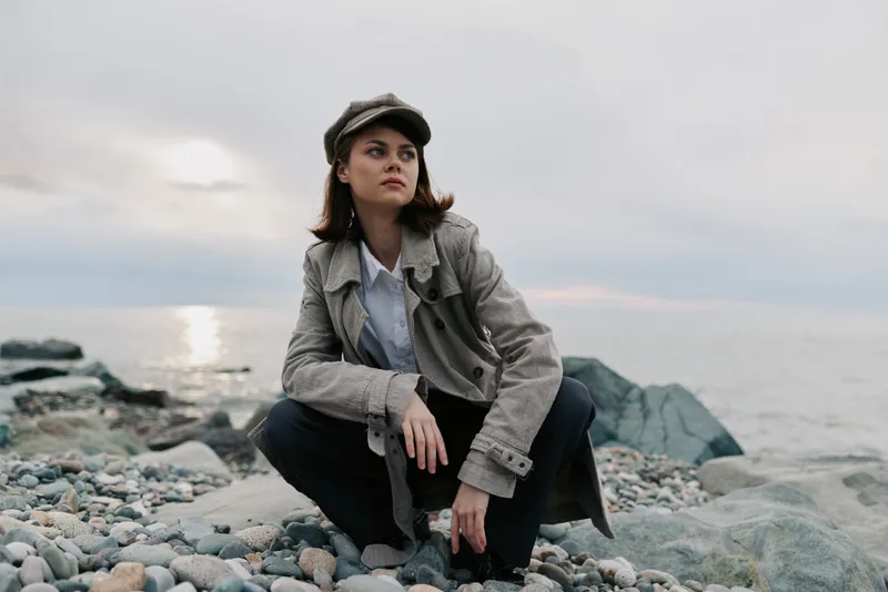 Young woman in grey coat and cap squats on rocky beach near calm sea at sunset. Moody natural light highlights thoughtful expression and casual autumn fashion by the water. Outdoor lifestyle.