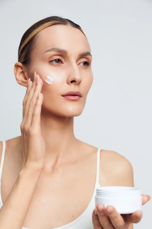 Young adult with blonde hair applies facial moisturizer to her face in a close up studio shot, capturing a calm skincare moment with soft lighting.