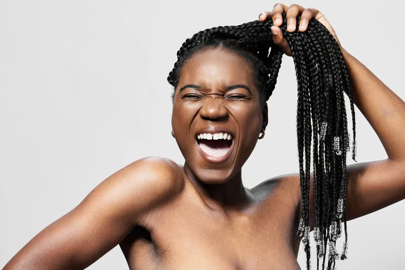Joyful young woman with braided hair laughing and playfully pulling her hair against a neutral background, showcasing a vibrant and confident personality