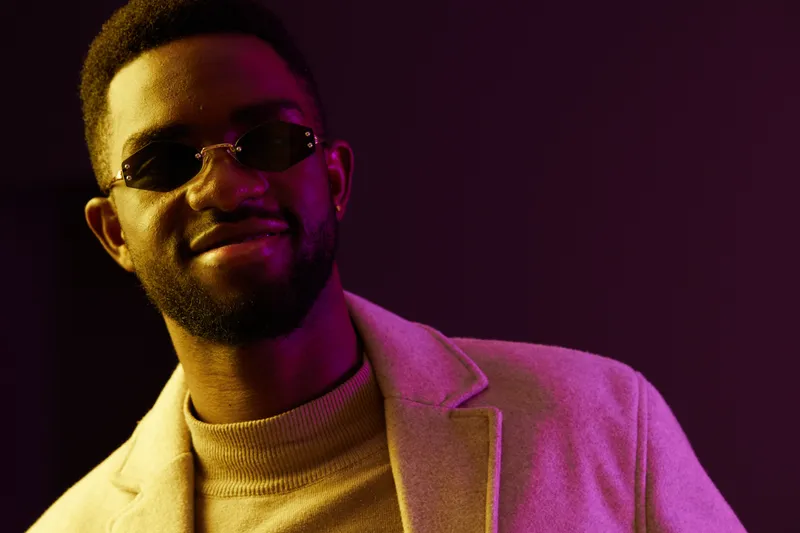 Trendy African man smiling confidently in stylish sunglasses and a light coat against a vibrant purple background Modern, youthful vibe for fashion