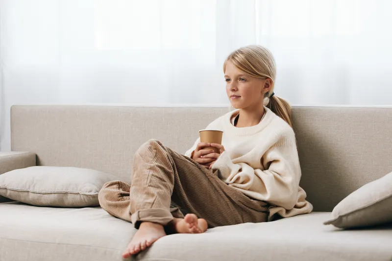 Child sits barefoot on a soft sofa, holding a paper cup, wearing a cozy sweater and relaxed pants. A calm, intimate moment of quiet home comfort and easy living.