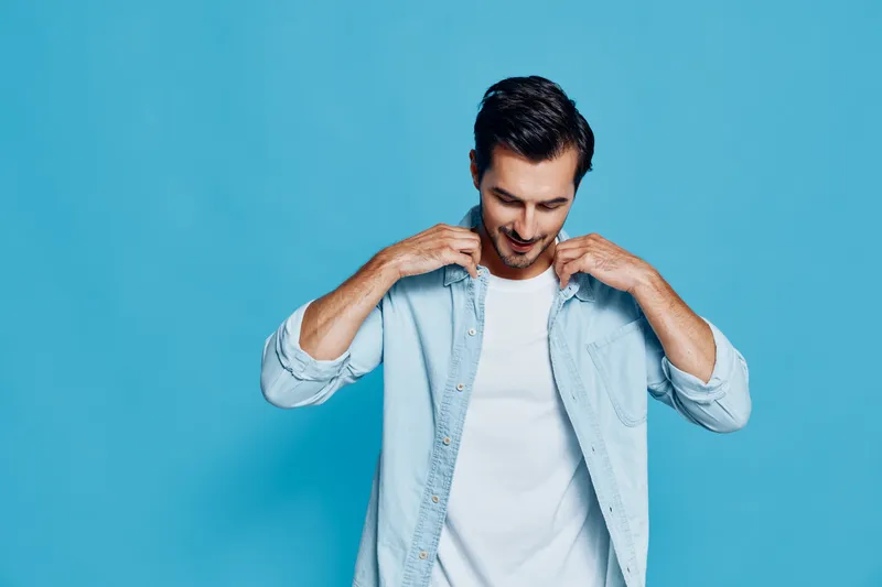 Stylish young man wearing a light denim shirt smiles while adjusting his collar against a vibrant blue background. Fashion concept and cheerful mood.