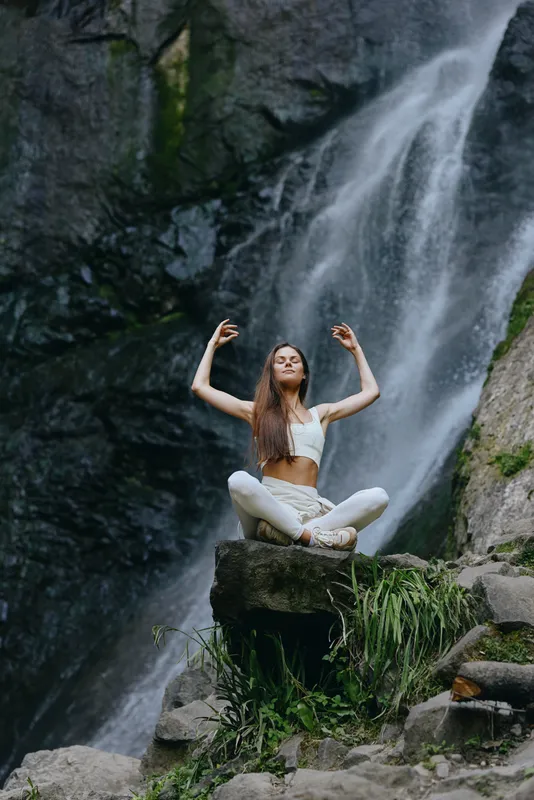 woman meditating outdoors near waterfall in white sportswear, serene nature scene with lush greenery and rocky terrain, peaceful yoga practice