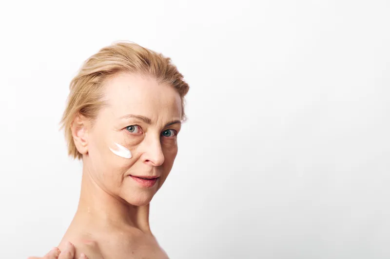 Mature woman with short blonde hair applies white face cream on cheek, looking at camera on white background. Concept of skincare, anti aging, natural beauty, and self care.