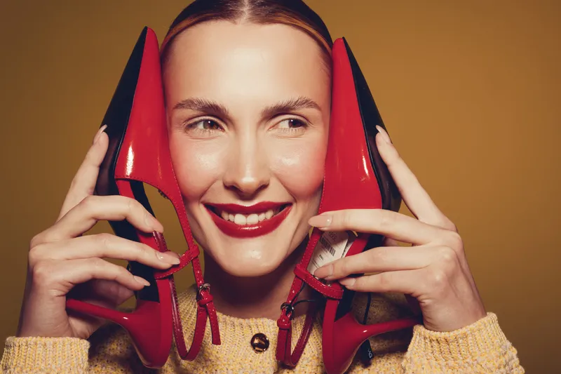 Smiling woman holds red high heels near face, wearing yellow sweater. Close-up portrait on brown background. Concept of fashion, style, and joyful mood with bright makeup.