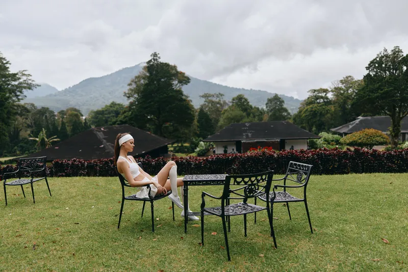 A serene outdoor scene featuring a female model in casual sportswear, sitting thoughtfully on a chair amid lush greenery, mountain backdrop, conveying calmness and fitness