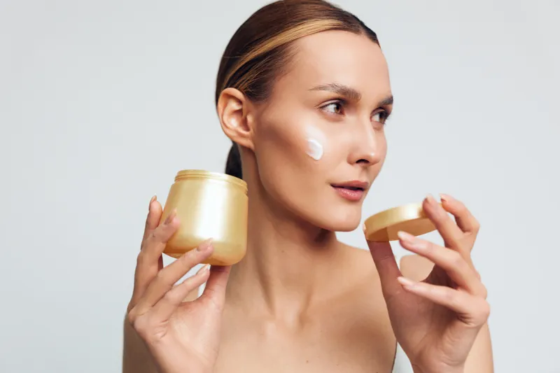 Skincare routine portrait of a woman applying moisturizer from a gold jar, clear skin, calm expression, cosmetic cream on cheek, studio shot with minimal background.