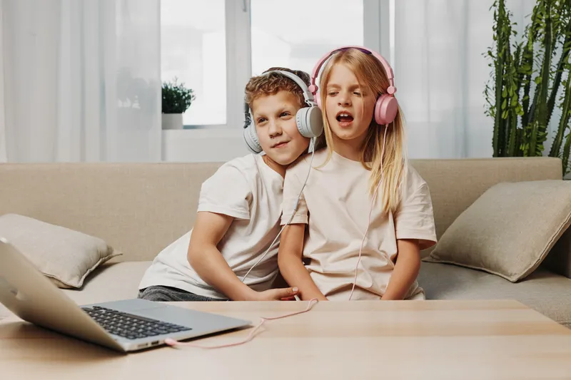Two happy children sitting on a couch wearing headphones and sharing music from a laptop in a bright living room. Concept of childhood, technology, and friendship.