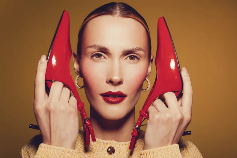 Portrait of a woman holding red high heel shoes near her face against a brown background. Fashion concept with bold makeup, stylish earrings, and confident expression in studio lighting.
