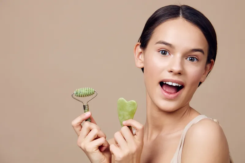 Young woman with a joyful expression holding a jade roller and gua sha stone, promoting skincare and self care routines in a neutral tone background