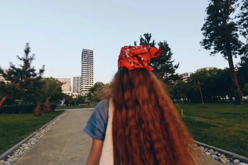 girl with long red hair wearing a red bandana walking in urban park surrounded by green trees and modern buildings under clear sky