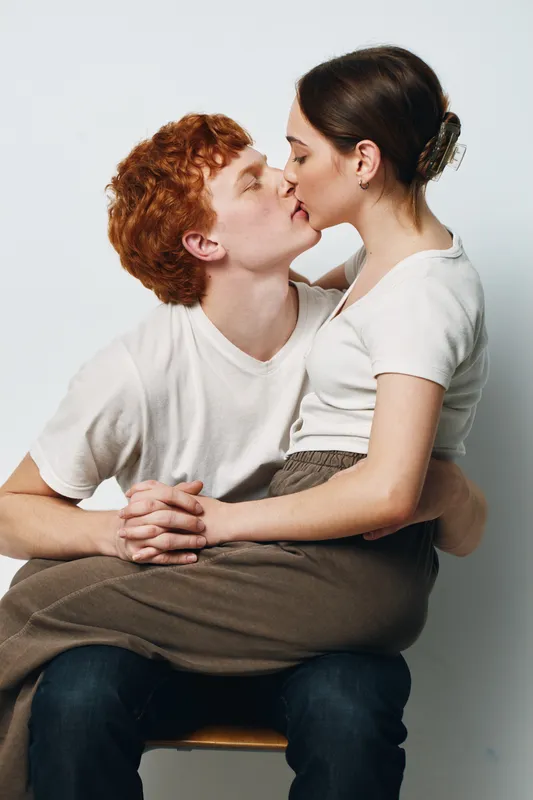 Couple kissing and holding hands, sitting close with affection and intimacy on white background. Young man and woman with casual clothes sharing romantic moment and connection.