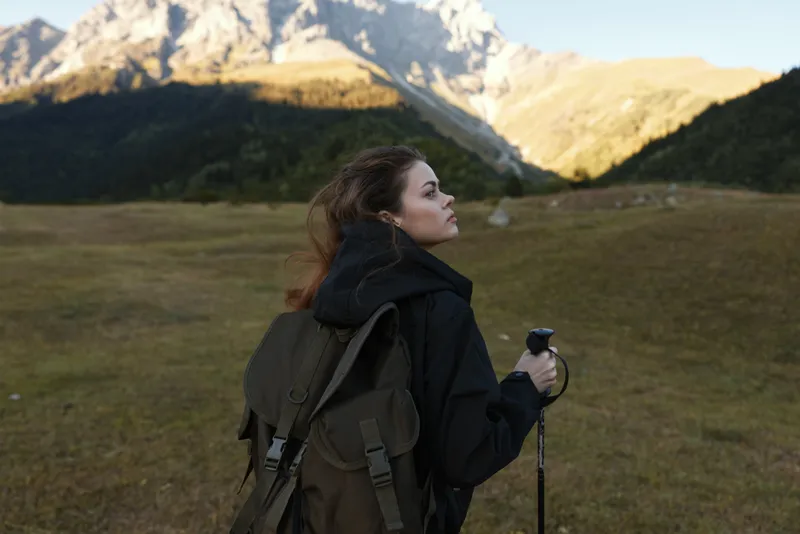 A lone female hiker stands with a backpack in a rugged mountain landscape, gazing toward distant peaks. She wears a dark jacket, ready for adventure, embodying calm focus and outdoor resilience.