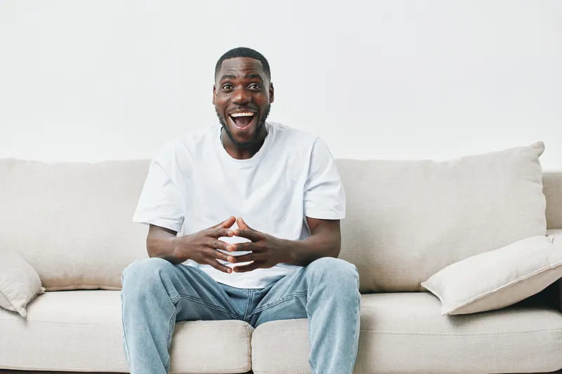 Happy young African man sitting on a couch, enjoying leisure time at home with a joyful expression, wearing casual clothing, showcasing comfort and relaxation
