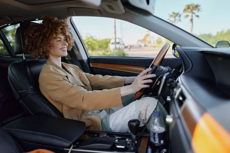 A woman behind the wheel in a stylish car, captured with retro film color and warm tone. She smiles as daylight pours through the window, conveying casual lifestyle and drive.