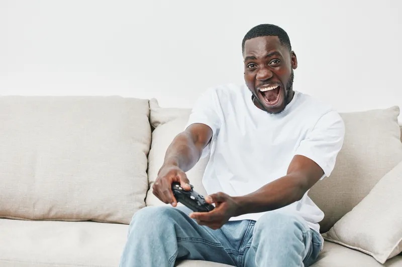 Excited African American man playing video games in casual white t shirt, showcasing joy and enthusiasm on lifestyle couch background