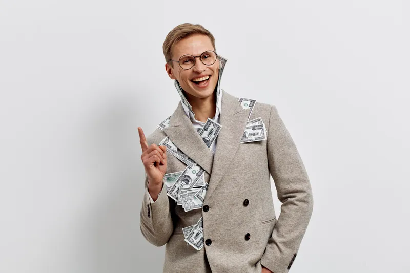 Smiling man in gray double-breasted suit with newspaper and money confetti, celebrating success, happy and confident expression, modern business attire, light background, studio portrait, positive emo