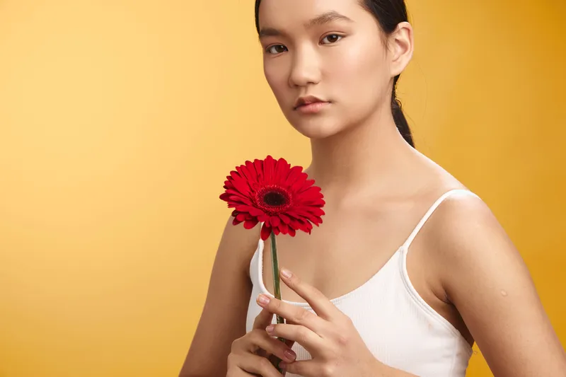 asian woman holding a red flower in a studio portrait, wearing a white tank top against a warm yellow backdrop, a calm expression conveying elegance and focus.