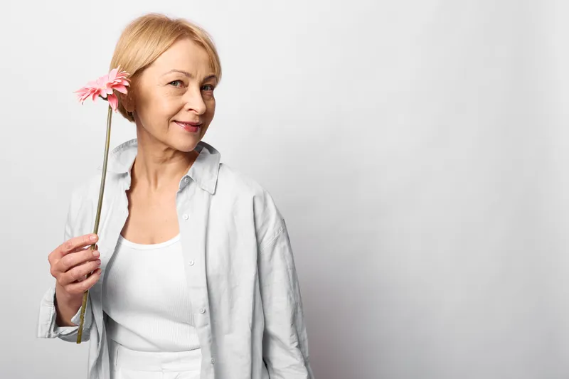 Elderly woman holding pink flower, gentle smile, relaxed pose, white studio background, soft natural light, short blond hair, calm spring mood, shallow focus, portrait, indoors