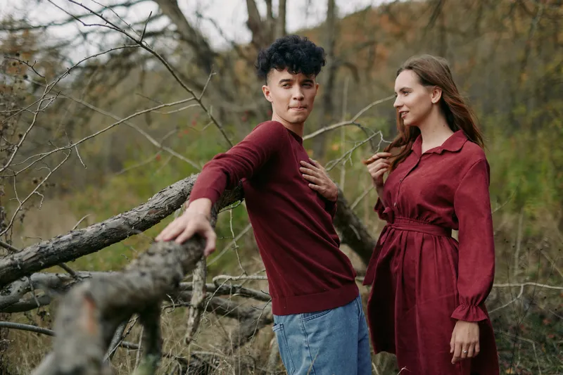 A candid scene features a young couple in earthy burgundy outfits outdoors, leaning on a fence with calm expressions, conveying genuine emotion and natural authenticity.