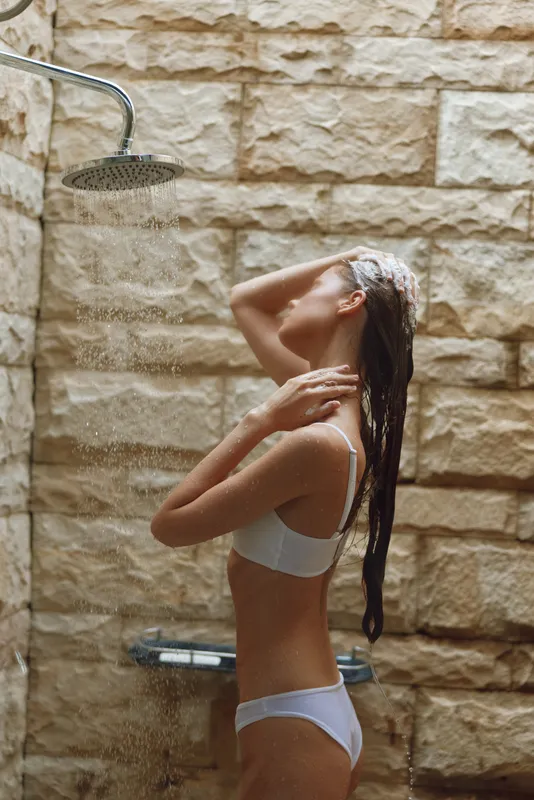young woman showering outdoors with wet hair, natural stone wall background, white underwear, relaxed mood in warm lighting, wellness and self-care concept