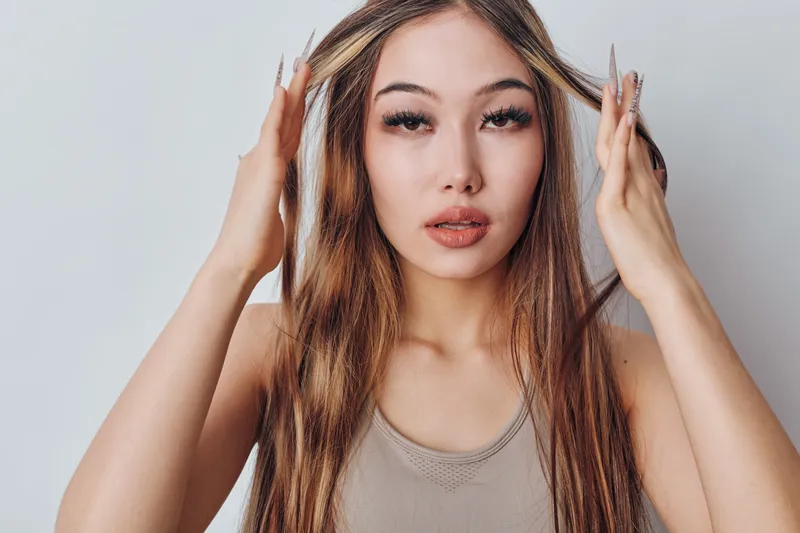 Portrait of young woman with long hair and makeup touching head with fingers, looking at camera on neutral background. Female model expressing calm and confidence in studio.