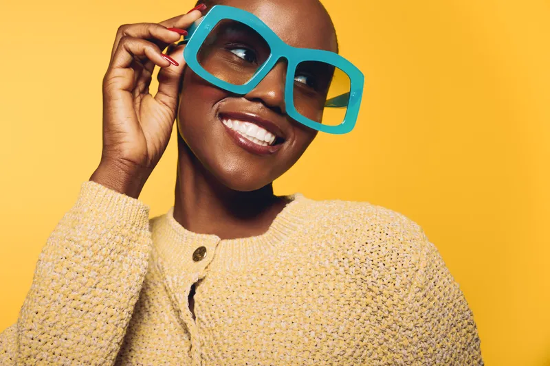 Smiling black woman with dark skin wearing large blue glasses and a beige knitted sweater poses against yellow background. Concept of fashion, style, happiness, and confident beauty.
