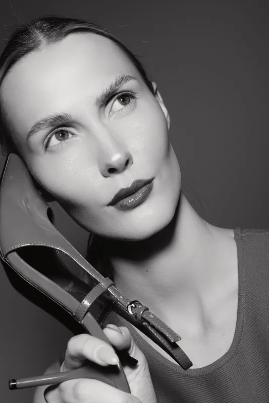 Portrait of a slender Caucasian woman with pale skin holding a high heel shoe and a pencil near her face. Studio lighting emphasizes her facial features and thoughtful expression.