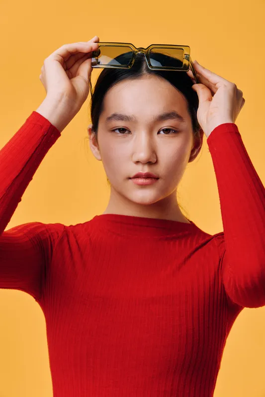 Young woman wearing red sweater putting on sunglasses against yellow background. Closeup portrait, fashion, casual style, confident expression, studio light, beauty and youth.