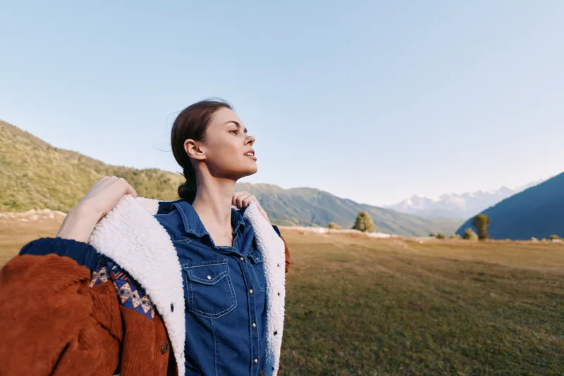 Woman portrait in mountains nature meadow wearing a coat and denim shirt, standing with eyes closed and enjoying fresh air, travel freedom and scenic alpine landscape relaxation.