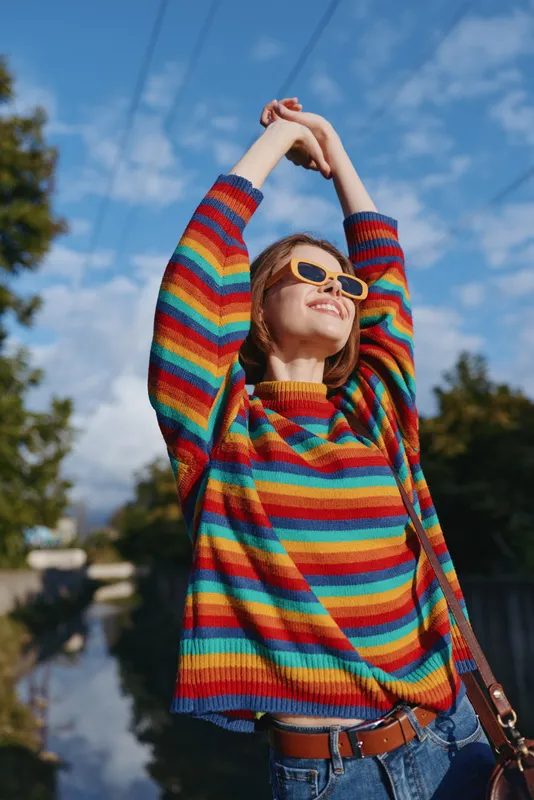 Young woman in a colorful striped sweater and yellow sunglasses smiling and stretching arms outdoors under blue sky, casual outfit, joyful lifestyle and urban nature backdrop.