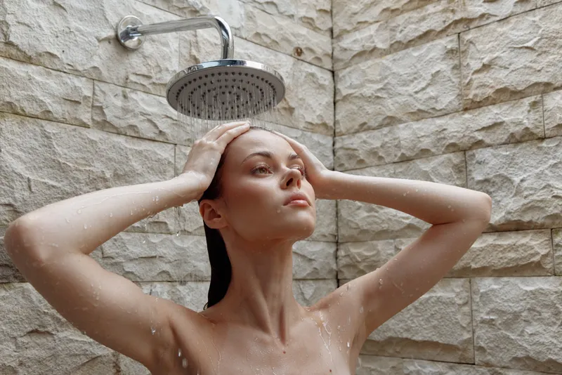 Woman washing hair under outdoor rainfall shower with stone wall background, natural light, serene expression, wellness and relaxation concept.