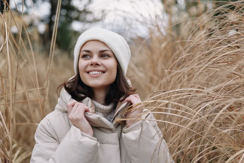 Woman in a puffy winter coat and knit hat smiles among tall dried grasses, warm scarf wrapped around her neck, on a light winter day in a tranquil natural outdoor portrait.