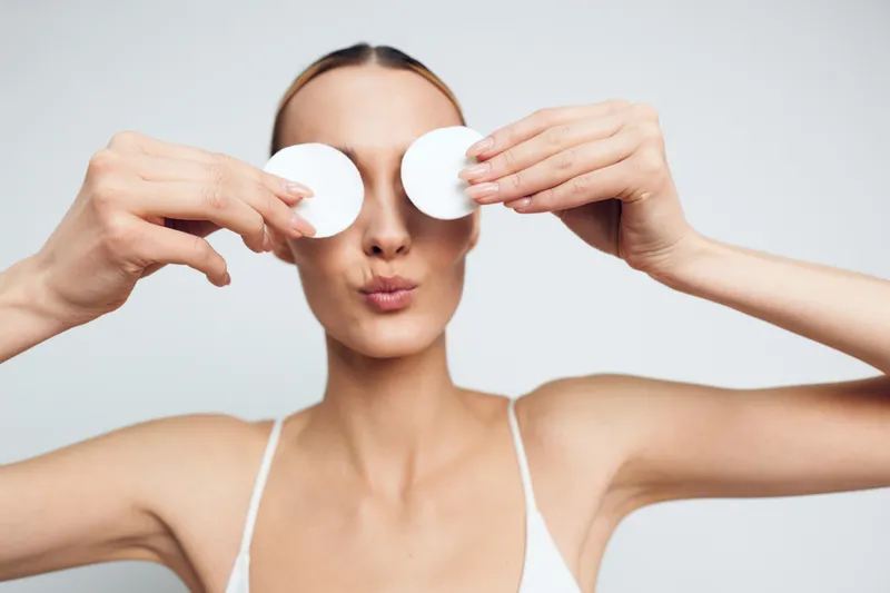 Young woman with clear skin holding cotton pads over eyes, skincare routine, beauty care, fresh face, white background, minimal makeup, clean and natural look