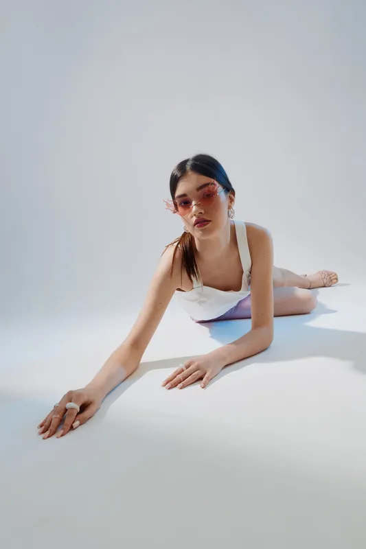 Young woman with long dark hair wearing pink sunglasses and white outfit lies on the floor in a bright studio. Concept of beauty, fashion, and modern style.