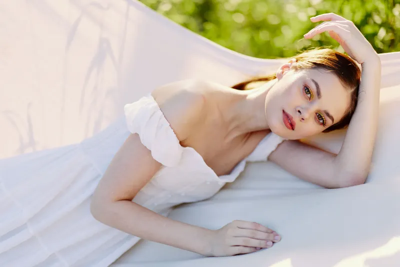 Beautiful young woman with soft features wearing a white dress, lying gracefully on a fabric in a lush green field, evoking tranquility and elegance during spring