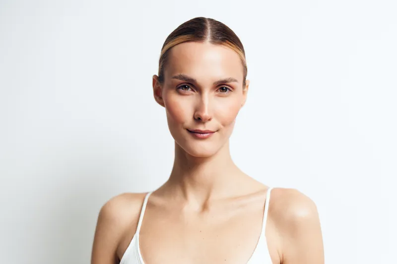 Portrait of a confident woman with smooth skin and pulled back hair wearing a white tank top. Minimalist white background highlights natural beauty and skincare concept.