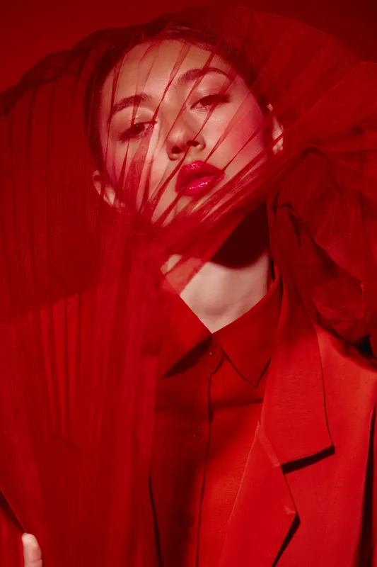 Fashion model with bold red lipstick and dramatic red tulle fabric around her face showcasing creative beauty styling and high fashion editorial concept.