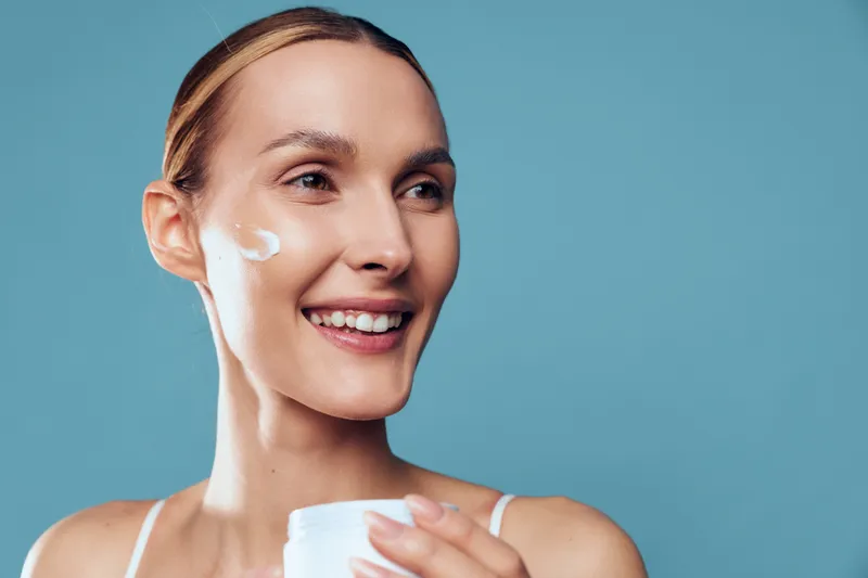 Smiling woman applying cream on face holding jar, natural skincare routine, healthy skin, clean beauty, closeup portrait, blue background, wellness, self care