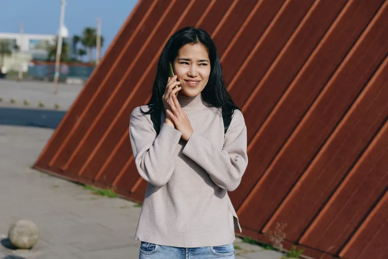 Woman talking on mobile outdoors in a light sweater and jeans beside a rusted wall, urban street scene, candid casual moment, cheerful expression.