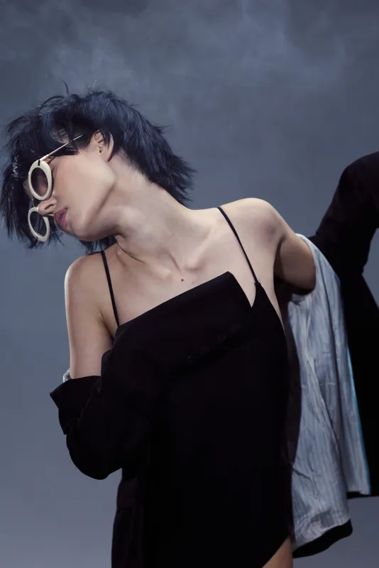 Stylish woman with short dark hair wearing sunglasses and a black dress in a moody studio setting