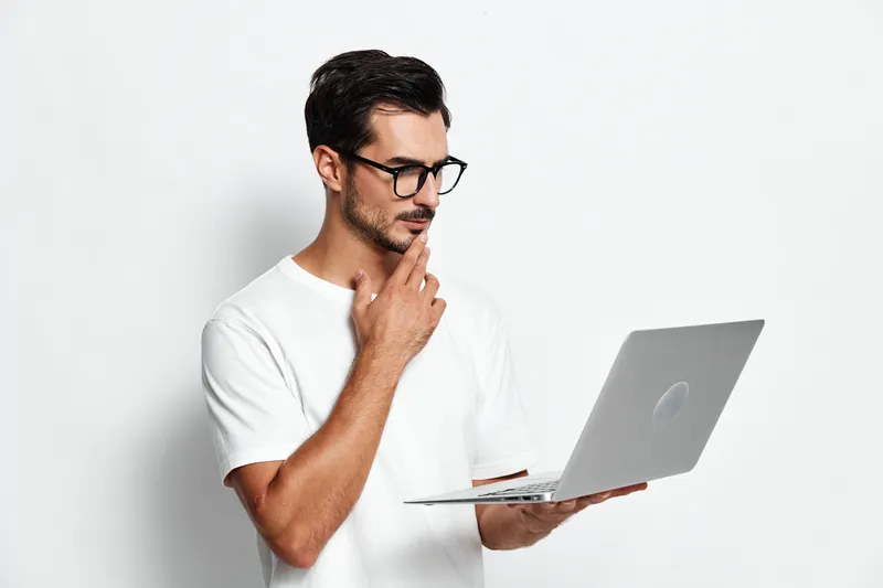Thoughtful young man in white t-shirt wearing glasses looking at laptop screen, pondering ideas with hand on chin, isolated on plain solid background. Professional concept.