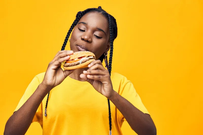 Young Black woman joyfully enjoying a delicious burger against a vibrant yellow background, highlighting the joy of healthy eating and fun food moments