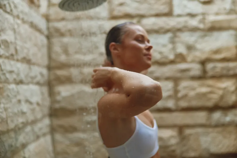 woman showering indoors with wet hair and relaxed expression under warm water spray in stone-tiled bathroom, calm and refreshing moment