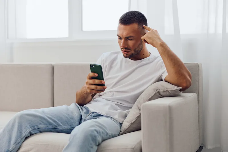 Thoughtful man sitting on a sofa, engaged with his smartphone, showcasing modern digital lifestyle and connectivity in a trendy, minimalist interior.