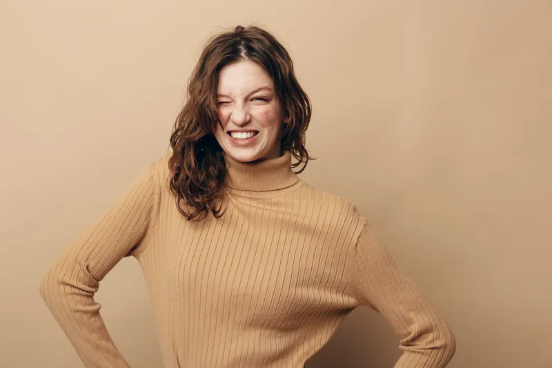 Young woman in beige sweater grimaces with eyes closed and hands on hips against neutral background. Concept of discomfort, frustration, and emotional expression in casual style.