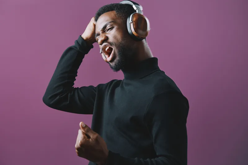 Young African man expressing passion while singing with headphones, against a vibrant purple background, showcasing emotions of joy and energy in a creative music concept