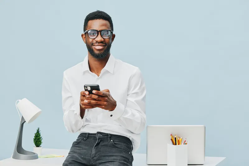 Smiling man with glasses dressed in white shirt using smartphone at modern workspace with white desk and laptop, cheerful professional in bright studio setting. People lifestyle concept