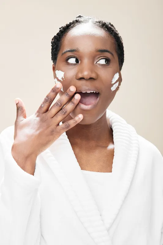 A surprised African woman applying skincare cream, showcasing a fresh and healthy beauty routine, with a neutral background and soft color palette